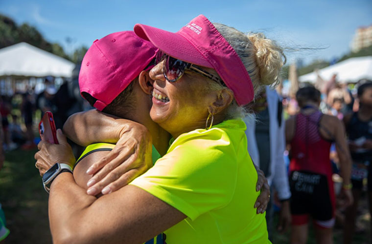 One woman wearing a pink hat and a yellow shirt hugs another woman wearing a pink visor and a yellow shirt at an athletic competition.
